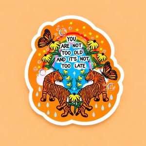 May include: A die-cut sticker with an orange background featuring two orange and black tigers facing each other. The design includes yellow flowers, monarch butterflies, a rainbow, bubbles, and raindrops. The text reads: YOU ARE NOT TOO OLD AND IT'S NOT TOO LATE.