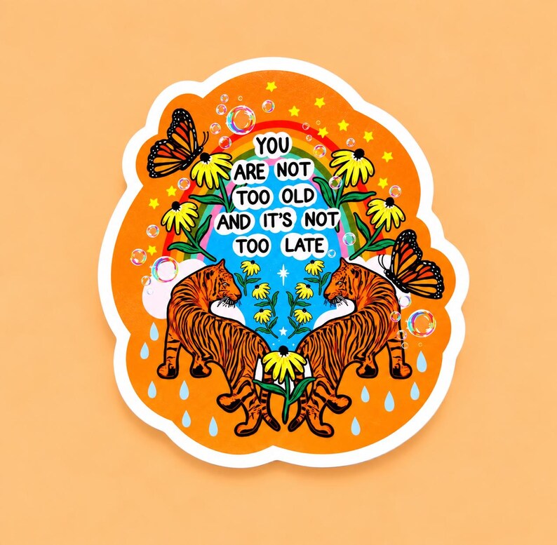 May include: A die-cut sticker with an orange background featuring two orange and black tigers facing each other. The design includes yellow flowers, monarch butterflies, a rainbow, bubbles, and raindrops. The text reads: YOU ARE NOT TOO OLD AND IT'S NOT TOO LATE.