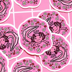 May include: Pink stickers featuring a cartoon axolotl with the text "LET ME BE WEIRD IN PEACE." The stickers are decorated with white daisies and red mushroom illustrations. The axolotl is pink with dark pink gills and a long tail.