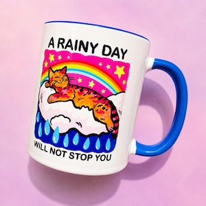 May include: A white ceramic mug with a blue handle and rim. The graphic features an orange cat resting on a white cloud under a rainbow and yellow stars. Below the cloud are blue raindrops. The text reads A RAINY DAY WILL NOT STOP YOU.