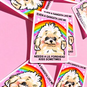 May include: A rectangular sticker featuring a cartoon illustration of a fluffy white dog against a rainbow background. The text reads EVEN A GANGSTA LIKE ME NEEDS A LIL FOREHEAD KISS SOMETIMES. The sticker has a pink border and white stars.