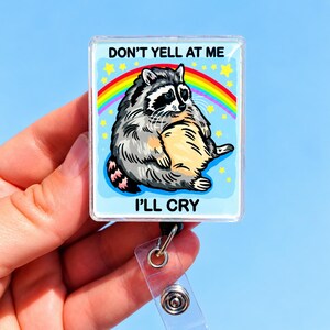 May include: A square badge reel featuring a cartoon illustration of a gray and black raccoon sitting in front of a rainbow and yellow stars. The text DON'T YELL AT ME is printed above the raccoon, and the text I'LL CRY is printed below it.