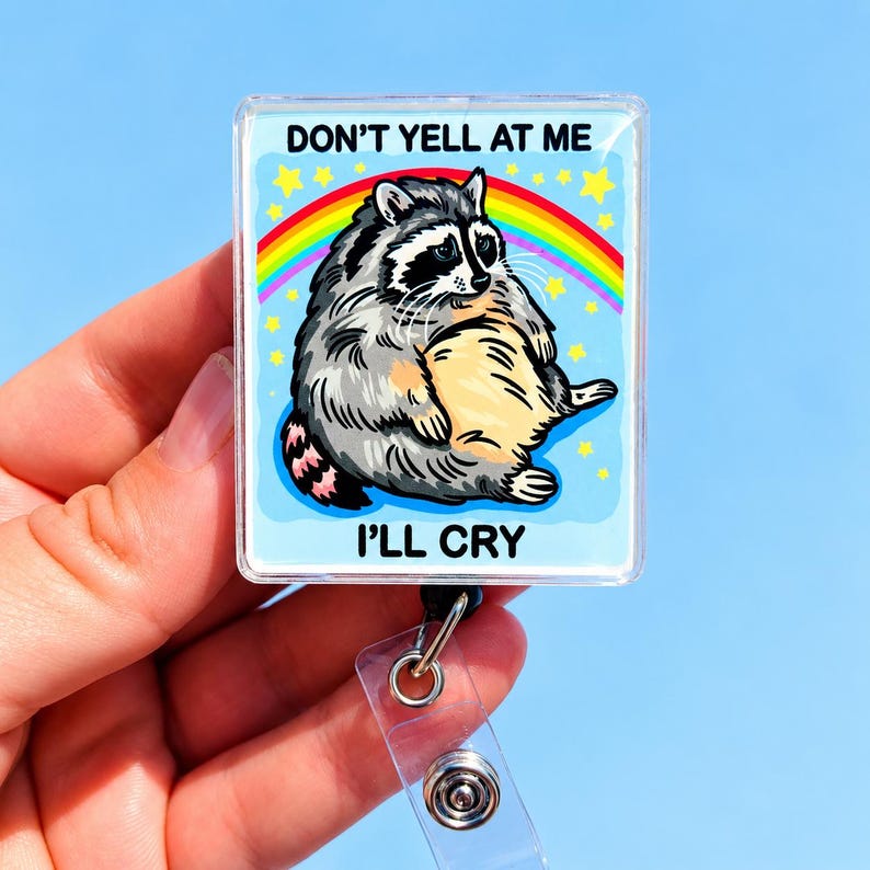 May include: A square badge reel featuring a cartoon illustration of a gray and black raccoon sitting in front of a rainbow and yellow stars. The text DON'T YELL AT ME is printed above the raccoon, and the text I'LL CRY is printed below it.