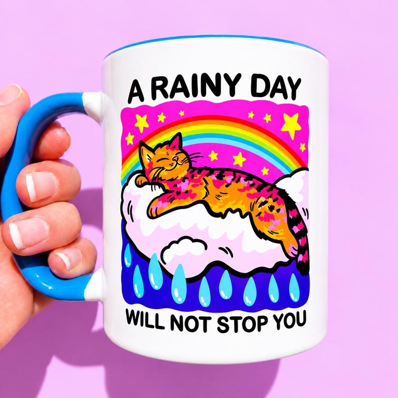May include: A white ceramic mug with a bright blue handle and rim. The graphic features an orange tabby cat sleeping on a white cloud under a rainbow and yellow stars, with blue raindrops below. Text reads A RAINY DAY WILL NOT STOP YOU.