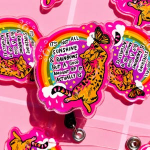 May include: A circular acrylic badge reel featuring an orange tabby cat reaching for a butterfly against a vibrant pink background with a rainbow, clouds, and the text: It's not all sunshine & rainbows but a good amount of it actually is.