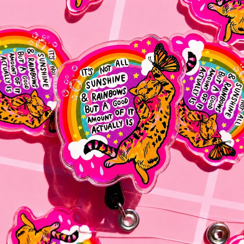 May include: A circular acrylic badge reel featuring an orange tabby cat reaching for a butterfly against a vibrant pink background with a rainbow, clouds, and the text: It's not all sunshine & rainbows but a good amount of it actually is.