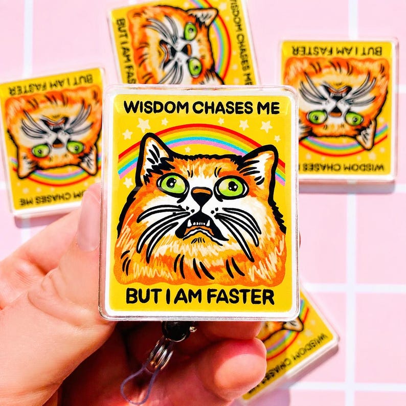 May include: A rectangular badge reel featuring a yellow background with a graphic of an orange cat with wide green eyes. Above the cat is the text WISDOM CHASES ME and below is the text BUT I AM FASTER. A rainbow arc and white stars are visible.