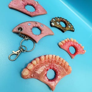 May include: A collection of five semi-circular objects with central holes, including one keychain. The items feature realistic gum-like textures, some with embedded human-like teeth, one with metallic studs, and one with small blue circular accents.