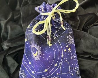 May include: A purple drawstring bag with a celestial print featuring stars, constellations, and a crescent moon. The bag has a yellow drawstring and purple beads.