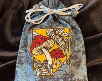 May include: A blue velvet drawstring bag with a white cord and an embroidered patch featuring two red and white mushrooms with black eyes on a yellow background.