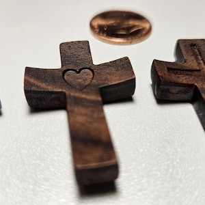 Könnte beinhalten: Nahaufnahme von drei kleinen, dunkelbraunen Holzkreuzen mit einem Cent-Stück. Ein Kreuz hat einen Herz-Ausschnitt, ein anderes ein geschnitztes Kreuz-Design, und das dritte hat das Wort "LOVE" eingraviert. Die Kreuze liegen auf einer weißen Oberfläche.
