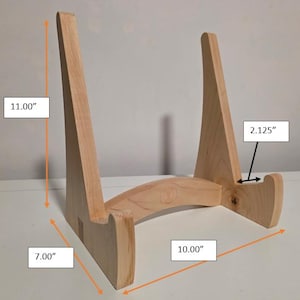 May include: A light-colored wooden display stand featuring two vertical supports and a curved base. The stand measures 11.00 inches in height, 10.00 inches in width, 7.00 inches in depth, and has a 2.125-inch ledge for holding items securely.