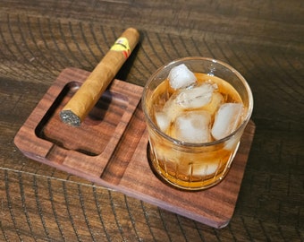 Op de afbeelding: Een houten sigarettenasbak met een sigaar en een glas whisky met ijs. De asbak is gemaakt van donker hout en heeft een rechthoekige vorm met een gedeelte voor de sigaar en een gedeelte voor het glas. Het glas is gevuld met amberkleurige vloeistof en ijsblokjes.