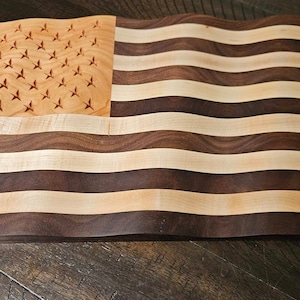 May include: A wooden cutting board shaped like the American flag. The board is made of walnut and maple wood and features a wavy design. The stars are burned into the maple wood.