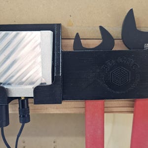 May include: A black 3D printed wall-mounted organizer with slots for tools. The organizer has a slot for a small electronic device with a cord, a slot for a wrench, and two slots for screwdrivers. The organizer has the text "3D Printing" and a 3D cube logo.