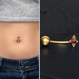 Puede incluir: Anillo de ombligo dorado con una piedra preciosa roja y una pequeña bola en el otro extremo.