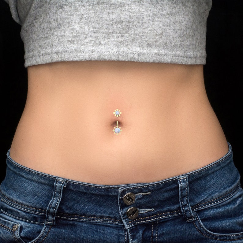 May include: A close-up of a person's midsection with a belly button ring. The ring is made of gold and has two small blue gemstones.