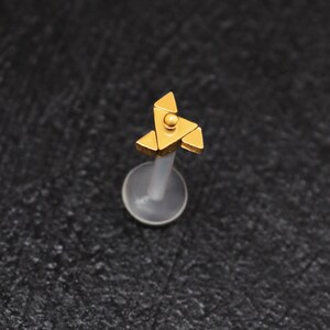 Puede incluir: Joyer&iacute;a corporal dorada con un dise&ntilde;o triangular. La joyer&iacute;a est&aacute; en un poste de pl&aacute;stico transparente.