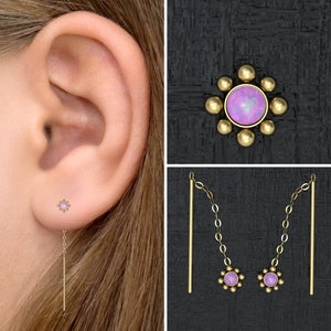 May include: A pair of gold earrings with a small pink opal gemstone in the center of each earring. The earrings are attached to a thin gold chain that hangs down from the opal.