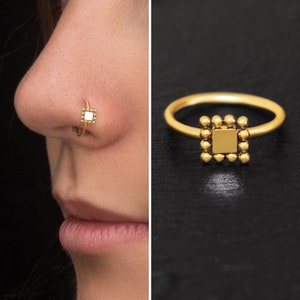 May include: A gold-colored nose ring with a square center and a ring with a square design. The nose ring is on a person's nose. The ring has a square design with small round accents.