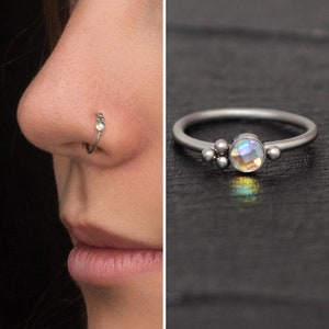 May include: A silver nose ring with a small iridescent gemstone and three small silver beads. The ring is shown on a person's nose and also laid flat on a dark surface.