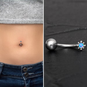 Puede incluir: Un piercing de ombligo plateado con un dije de flor de ópalo azul. El anillo es curvo y tiene una pequeña bola en un extremo.