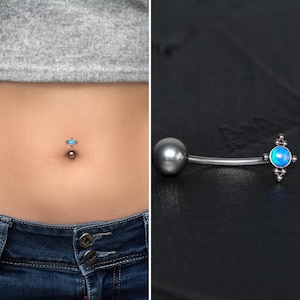 Puede incluir: Un anillo de ombligo plateado con una piedra preciosa de ópalo azul. El anillo se muestra en el ombligo de una persona y también se coloca sobre una superficie negra.