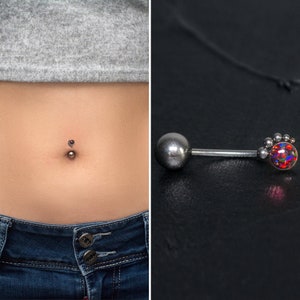 May include: A silver belly button ring with a small, clear gem in the center. The ring is shown on a person's belly button and also on a black surface.