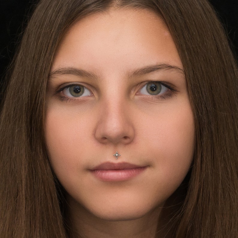 May include: Close-up portrait of a person with long brown hair. The person has a small, silver stud piercing in their lower lip.