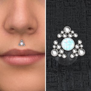 Peut inclure: Un bijou de corps en argent avec un groupe de petites perles argentées entourant une pierre plus grande ressemblant à une opale. Le bijou est présenté sur la lèvre d'un modèle.