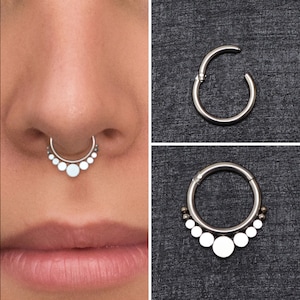 May include: A silver septum ring with a row of small white beads. The ring is shown on a person's nose and also on a black background.