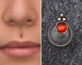 May include: A red gem with a silver top and three silver balls is attached to a clear barbell. The barbell is worn as a lip piercing.