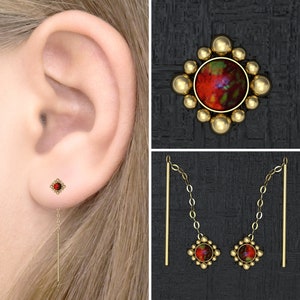 May include: A pair of gold earrings with a red gemstone and a dangling chain. The earrings are shown on a model's ear.