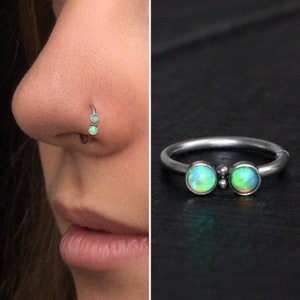 Può includere: Un anello per il naso in argento con due pietre di opale. L'anello è mostrato sul naso di una persona e anche su una superficie nera.