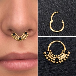 Peut inclure: Anneau de septum en or avec trois perles noires et un motif décoratif. L'anneau est présenté sur le nez d'un mannequin.