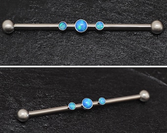 Puede incluir: Una barra de plata con tres gemas de ópalo azul. La barra es una joya de piercing corporal.