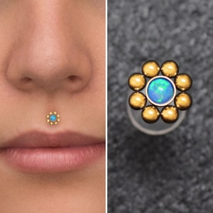 Puede incluir: Una joya de cuerpo de oro en forma de flor con una piedra preciosa de ópalo azul en el centro. La joya se muestra en un piercing en el labio inferior de una modelo.