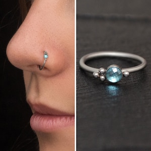 Könnte beinhalten: Ein silberner Nasenring mit einem kleinen, hellblauen Edelstein und drei kleineren silbernen Perlen. Der Ring wird an der Nase einer Person gezeigt und auch separat auf einer dunklen Oberfläche dargestellt.