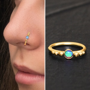 Puede incluir: Anillo de nariz de oro con una pequeña piedra de ópalo iridiscente y un diseño de cuentas.