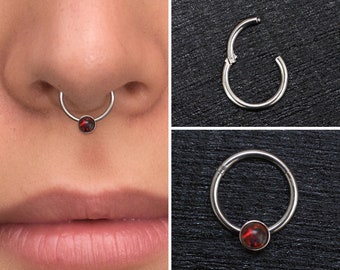 May include: A silver hinged segment ring with a red opal gemstone. The ring is shown in a close-up view and also being worn in a nose piercing.