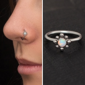 Può includere: Un anello per il naso in argento con un piccolo opale bianco incastonato in un design floreale. L'anello è mostrato sul naso di una persona.