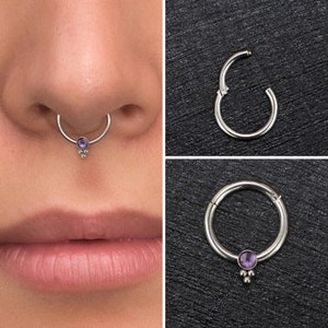 Puede incluir: Un anillo de septum de plata con una pequeña piedra preciosa púrpura y tres pequeñas cuentas de plata que cuelgan de él. El anillo se muestra en la nariz de una persona.