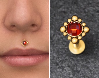 May include: A gold-colored lip piercing with a square, red gemstone center and small, round gold accents. The piercing is shown in use on a person's lip and also displayed separately against a dark background. Jewelry.