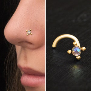 Puede incluir: Anillo de nariz de oro con una piedra preciosa transparente iridiscente y cuatro pequeñas cuentas doradas. El anillo se muestra en la nariz de una modelo y también sobre una superficie negra.