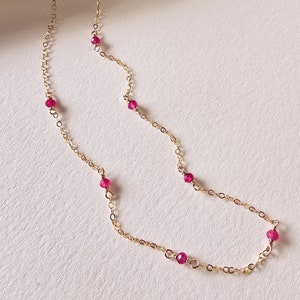 Op de afbeelding: Een delicate gouden ketting met kleine, felroze edelstenen. De ketting ligt op een wit oppervlak.