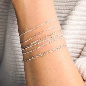 Puede incluir: Primer plano de una muñeca con varios brazaletes de cadena plateados. Los brazaletes están superpuestos, creando un look elegante y delicado.