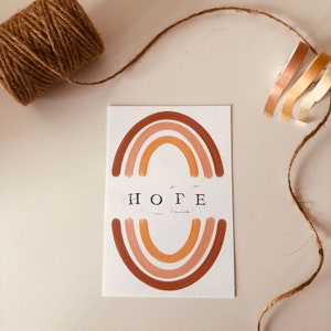 May include: A white card with a brown, orange, and pink rainbow painted on it. The word "HOPE" is printed in black letters below the rainbow.