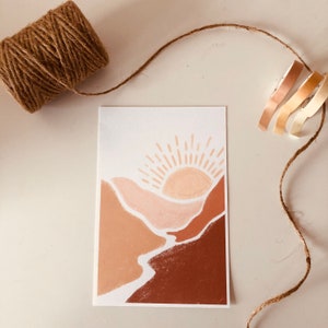 May include: A minimalist art print featuring a stylized sunset over a landscape of brown, beige, and pink hills. The sun is depicted as a simple circle with rays emanating from it.