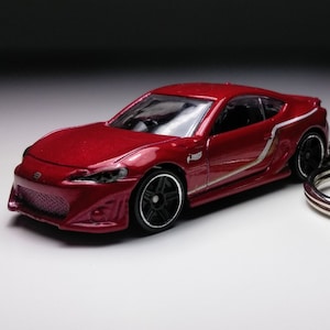 May include: Red toy car keychain with white racing stripes. The car is a replica of a Toyota 86.