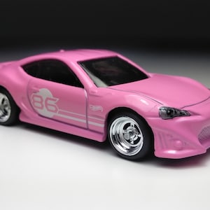 May include: Pink toy car with a white number 86 on the side. The car has a black grill and black wheels.